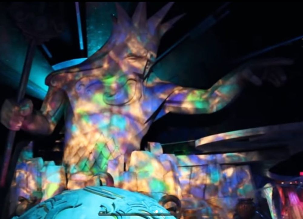 A large statue depicting the sea god Neptune looms over the photographer. The sculpture is of a man with a long beard and hair, no shirt, with a trident in one and while the other is outstretched further into the room. While the sculpture is made of white material, colorful lights are currently projected onto it in shades of purple, green, blue, and yellow.