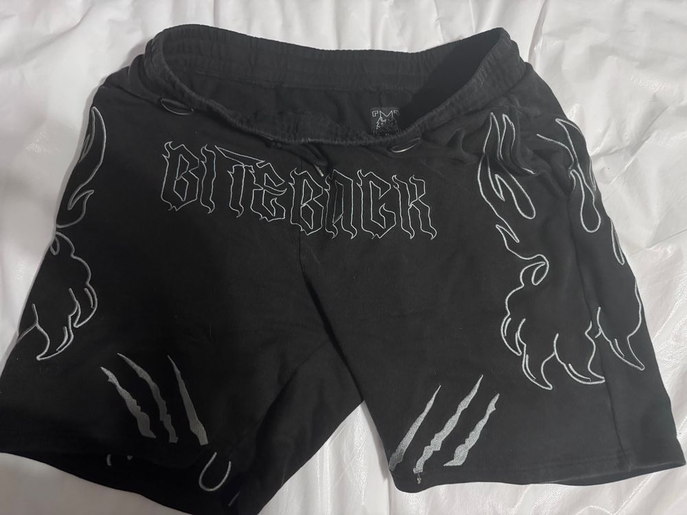 A pair of black shorts. On the front center reads “BITEBACK”. There are two stylized flaming clawed paws on the outside, and claw marks on the lower inside. The shorts also have metal loops in a few different places, which is unseen in the picture. All the designs are in silver.