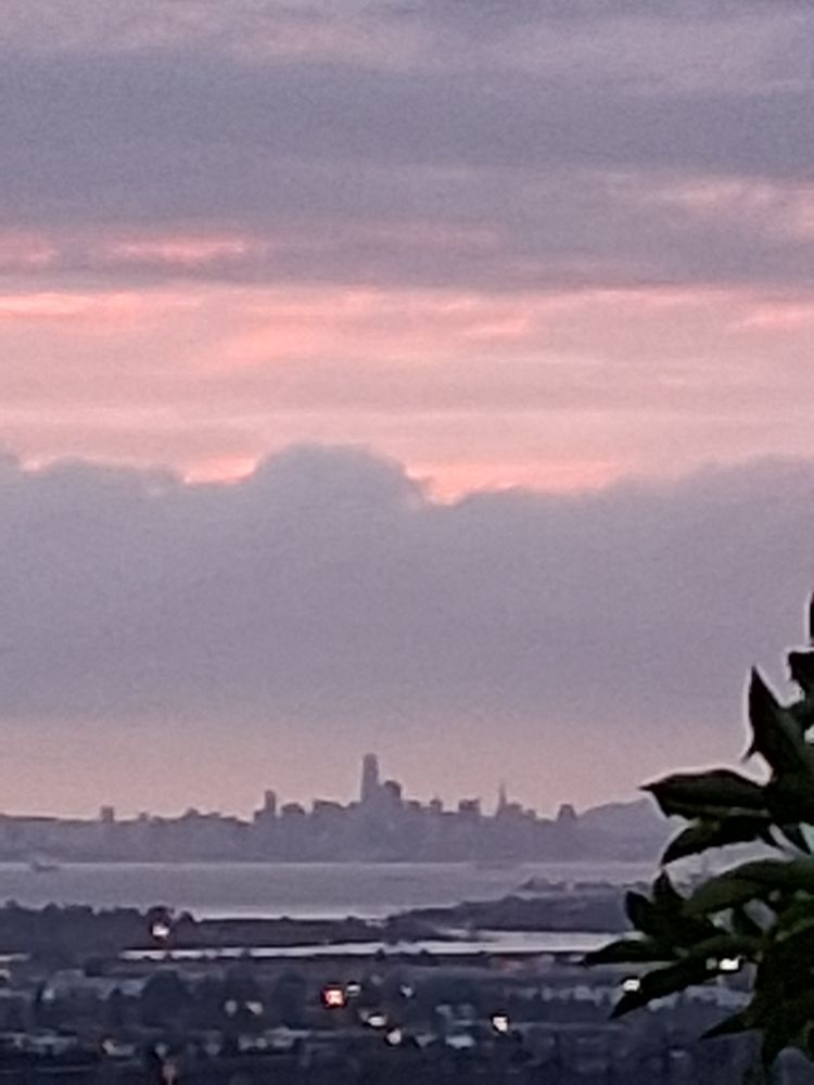 San Francisco skyline from the east, across the bay. Gray skyline topped with gray and pink clouds, pearly light from behind the  cliudbank. 