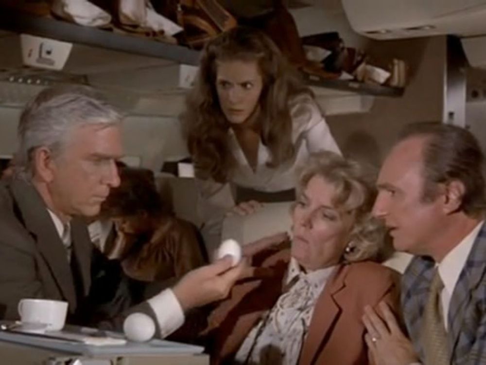 The egg scene from the movie Airplane! with Dr Rumack (Leslie Nielsen) kneeling in front of the passenger Shirley (Mary Mercier) holding an egg in his right hand that he has just removed from her mouth. The stewardess Elaine (Julie Hagarty) is looking directly at the egg with a concerned look. Shirley's husband is to her left comforting her.