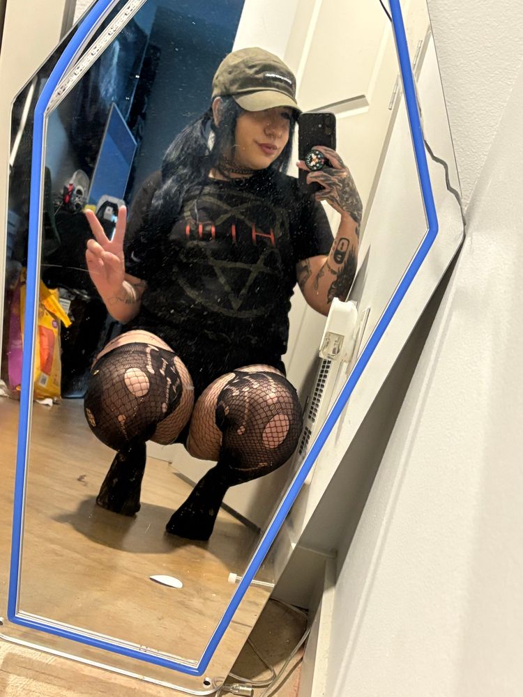Prinxe is squatting in front of a coffin mirror, wearing ripped fishnets and thigh highs. They have a HIM band tee on and small booty shorts to show off their thick thighs 