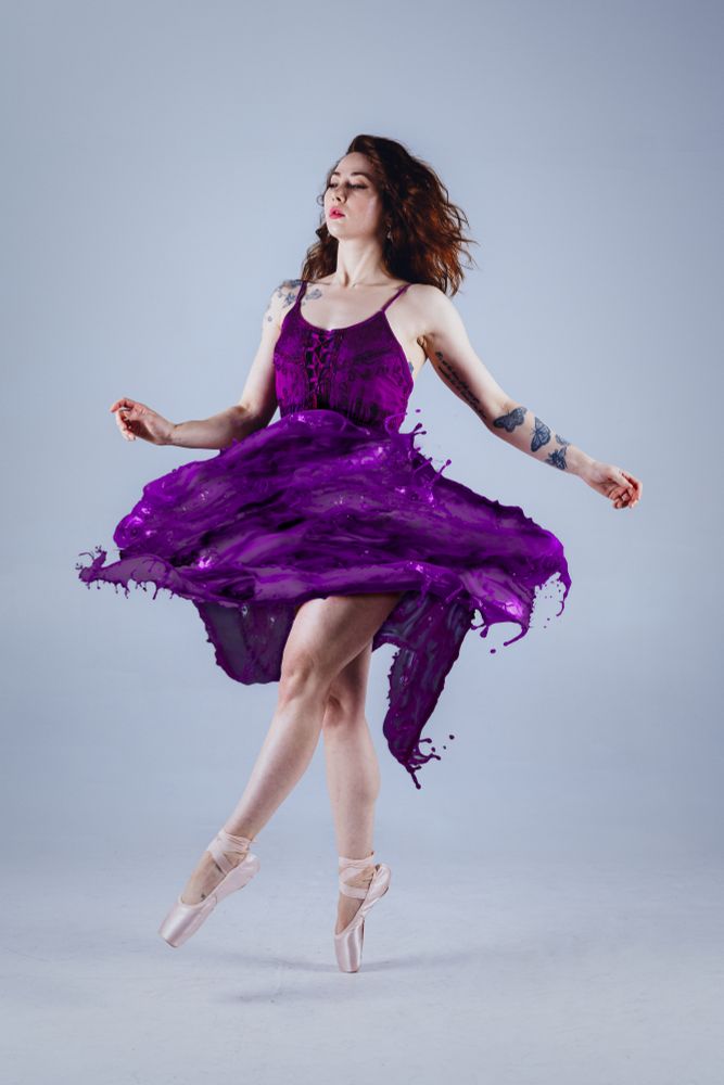 A dancer / model stands on pointe in ballet shoes. Wearing a deep purple dress where the skirt has been replaced to look like it's made of water swirling around them instead of fabric.