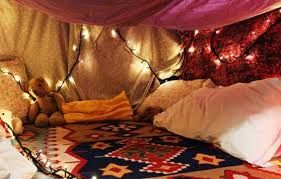 Cosy pillow fort with a multicoloured rug, large pillows, pink, red and white sheets for the walls and roof, lit up with warm white fairy lights.