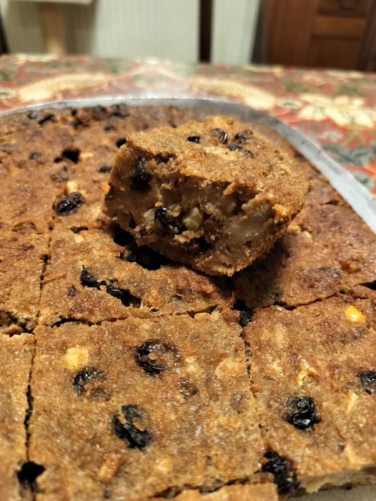 A slice of bread pudding on top of a dish of more slices. Predominantly brown, with lots of raisins and mixed peel visible. Not custardy!