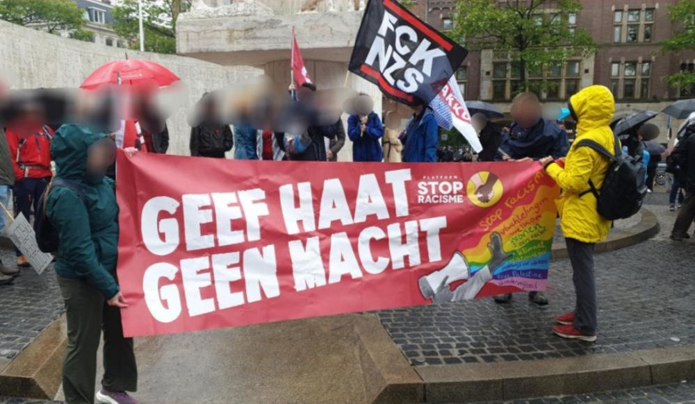 Regenachtige foto van het spandoek 'GEEF HAAT GEEN MACHT' van Platform Stop Racisme en Fascisme op de Dam. Op de achtergrond zie je onder andere een FCK NZS (Fuck Nazis) vlag.