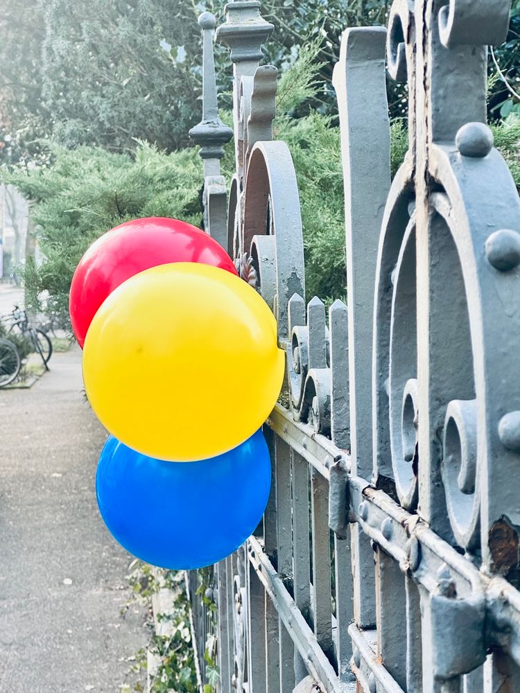 In this scene, there is a metal fence with ornate designs. Attached to the fence are three colorful balloons: one red, one yellow, and one blue. The balloons are arranged in a vertical line, with the red balloon at the top, followed by the yellow, and the blue at the bottom. Behind the fence, there are trees and bushes, suggesting a park or garden setting. A bicycle is parked nearby on the pathway.