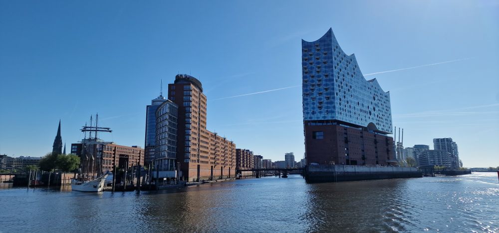 Aufnahme von einer Fähre auf der Elbe aus. Rechts die Elbphilharmonie, links das Columbus-Haus, davor ein Segelschiff, dahinter die Speicherstadt. Wolkenloser blauer Himmel