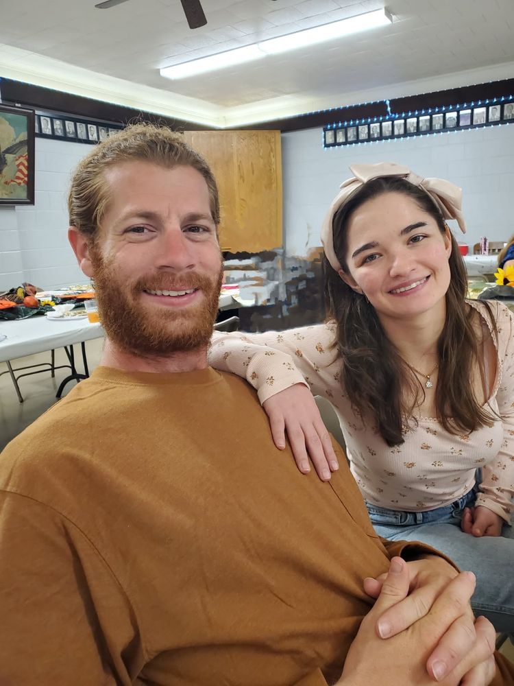 Red headed guy with orange shirt smiles next to brunette girl in pink floral shirt. 