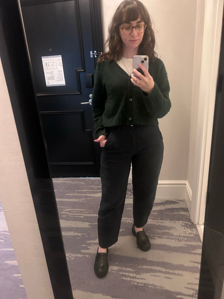 An image of me (I’m a white woman with long brown hair and glasses) wearing dark blue trousers, a white t shirt and a dark green wool cardigan buttoned, and black leather loafers 