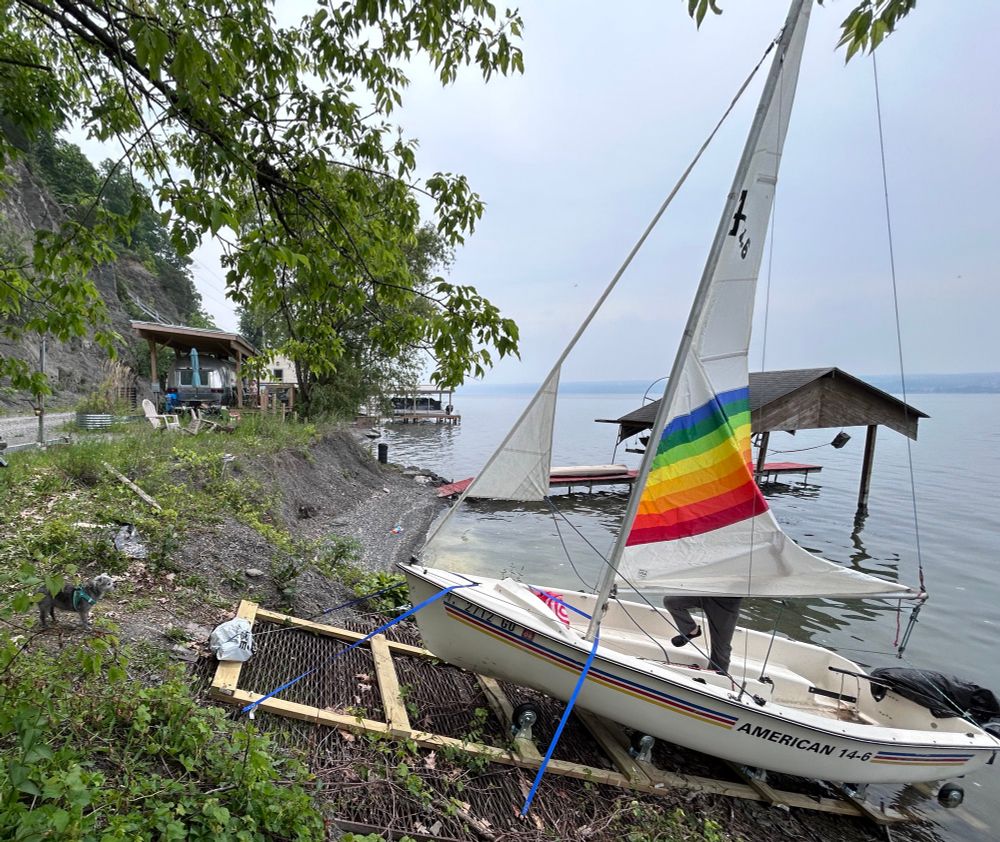 Image of a small 14 foot sailboat (AMERICAN 14-6) on a ramp on a lake. The sail has a rainbow color. 