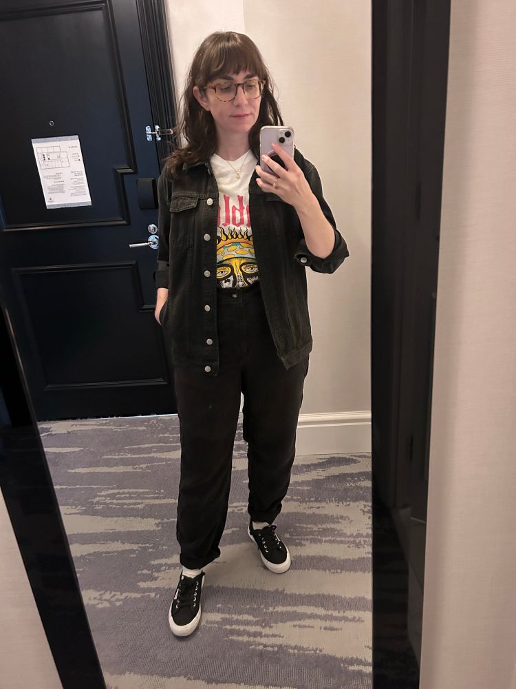 An image of me (I’m a white woman with long brown hair and glasses) wearing a sublime t shirt, a dark denim jacket, black trousers, and black superga shoes 