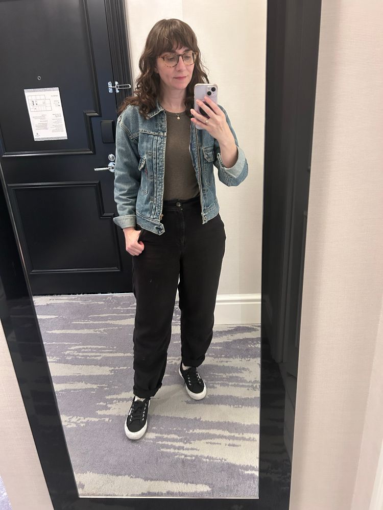 An image of me (I’m a white woman with long brown hair and glasses) wearing a gray tank top with a blue jean jacket on top, black trousers, and black superga shoes 