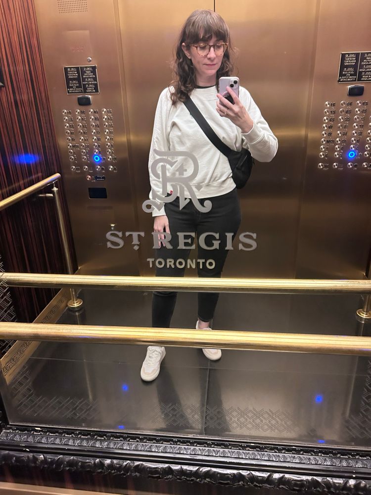 An image of me (I’m a white woman with long brown hair and glasses) wearing a white sweatshirt, dark gray jeans, and white sneakers. I’m in an elevator 