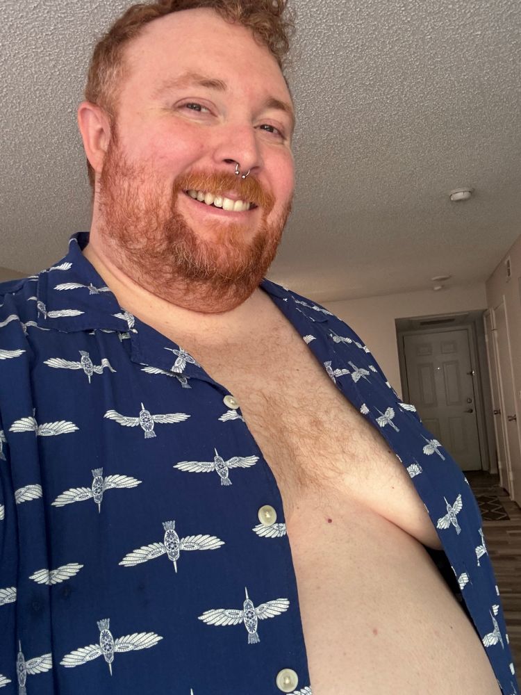 Cooper smiling at the camera with his blue button-up shirt open, reveal some tasteful cleavage