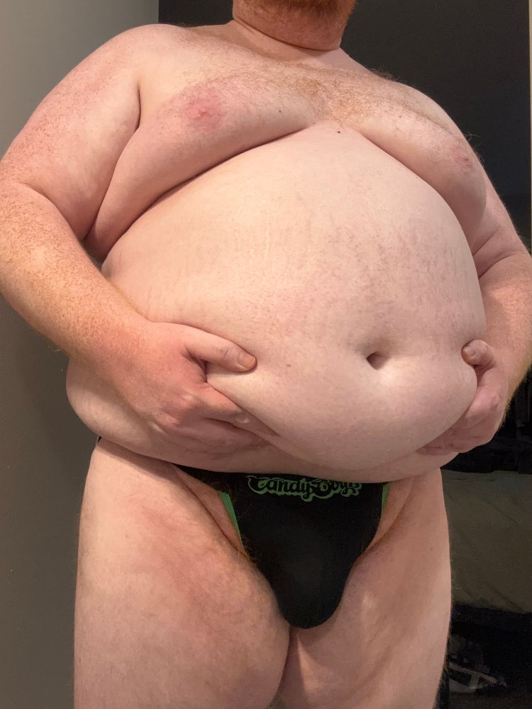 Cooper in a tight jock exposing his fat pad. He’s holding up his big round belly