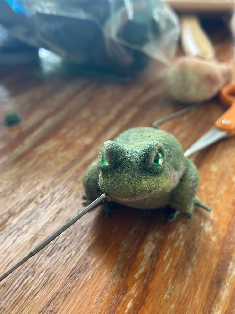 Front view of green frog (probably more of a toad) with bright green eyes, made from needle felted wool. He’s unfinished, still lacking front legs. 