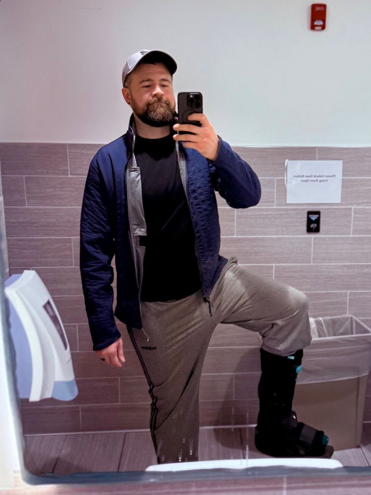 Bathroom selfie of bearded man in blue jacket and grey track pants holding up one leg showing the bottom half encased in a walking boot