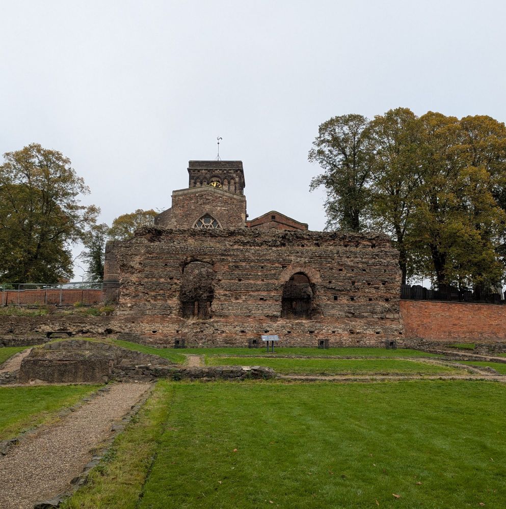 A Roman wall, quite high, with two arch windows. Behind is the square tower of a church and trees. Grass in front and some ground level Roman remains. Overcast sky 