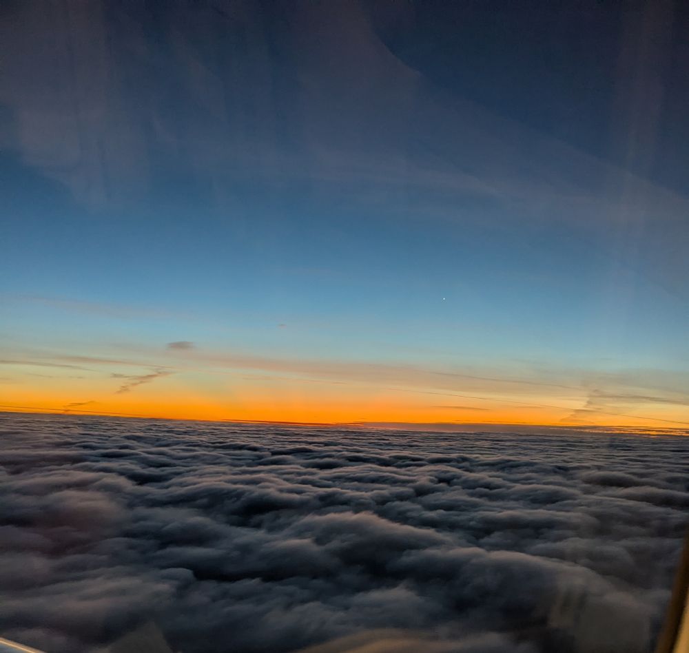 View from a plane window above the clouds. The clouds are dark and look almost like waves. Above, a stretch of pinkish orange from the dawn and blue above