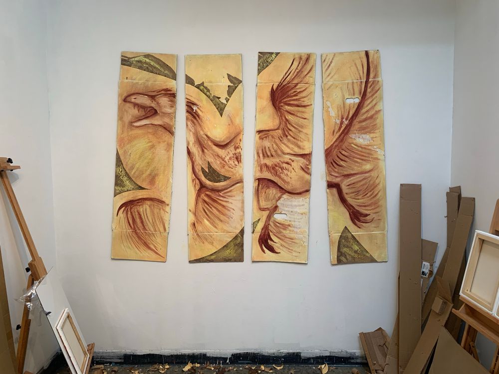 A dimly lit, small, cluttered artist studio with a painting on the wall. It’s fragmented into four panels from a cardboard box, depicting a reddish brown outline of a microraptor on a bright yellow-cream background. The cardboard has been slashed and dug into revealing the inner texture in multiple areas.  The pose is agonized and warped, filling a role between cave painting and imprint. 
