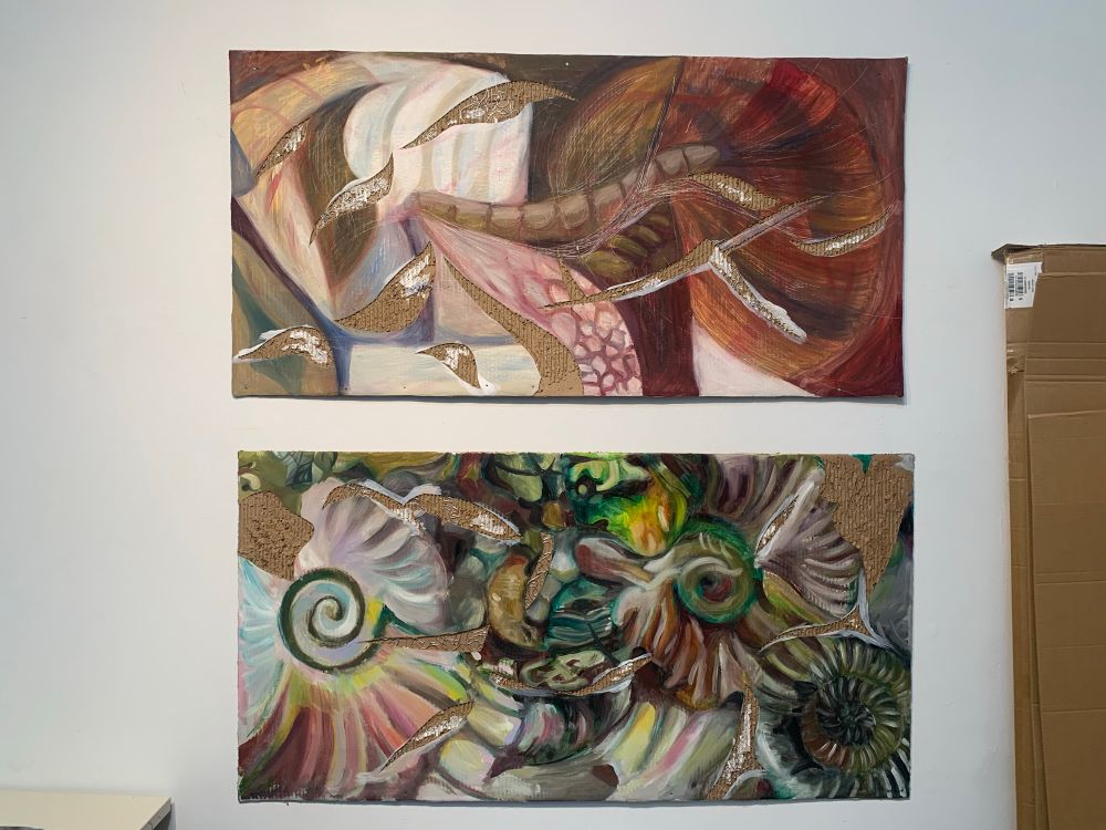 Two paintings hanging on a wall one on top of the other with a 2 inch gap between them. They’re both oil paint over top cardboard. The top painting is referencing prehistoric plants and the bottom one, prehistoric shelled animals such as ammonites. They read as very abstract. The top one is brown and orange with bits of scale texture and blue. The bottom is rainbow at times with multiple swirls and complicated clusters of form. Both have been cut into, revealing the textured cardboard underneath. In some of these instances, aluminum wire is decoratively woven throughout. 