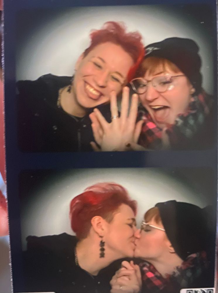 Two of a strip of photobooth pictures, with Simon and Chloe. In the top picture, Simon holds up their left hand to show off a silver band and grins, tongue poking out slightly. Chloe is excitedly smiling and pointing at the ring.
In the bottom picture, they have grabbed hands to kiss. 