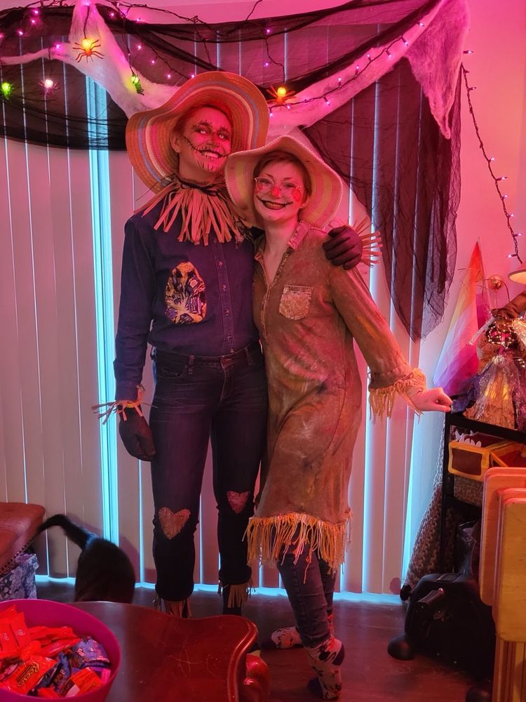 Two people dressed as scarecrows stand with arms around each other, grinning. The left one, Simon, is taller, with patched denim shirt and pants with faux straw at the cuffs and collar. The right one, Chloe, wears a long shirt dress covered in paint smears, with hay-looking yarn sewn around the hem and shirt cuffs. Both have large floppy straw hats and “scarecrow” makeup: stitched mouths and noses. They are standing in a home, with a large window and blinds behind covered in string lights and cobwebs.