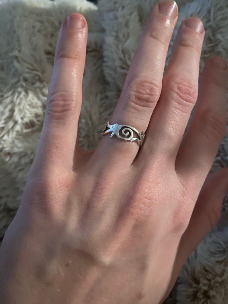 Close up of Simon’s hand, wearing the engagement ring. It is a silver band that looks like connecting spirals, without any gemstones. 