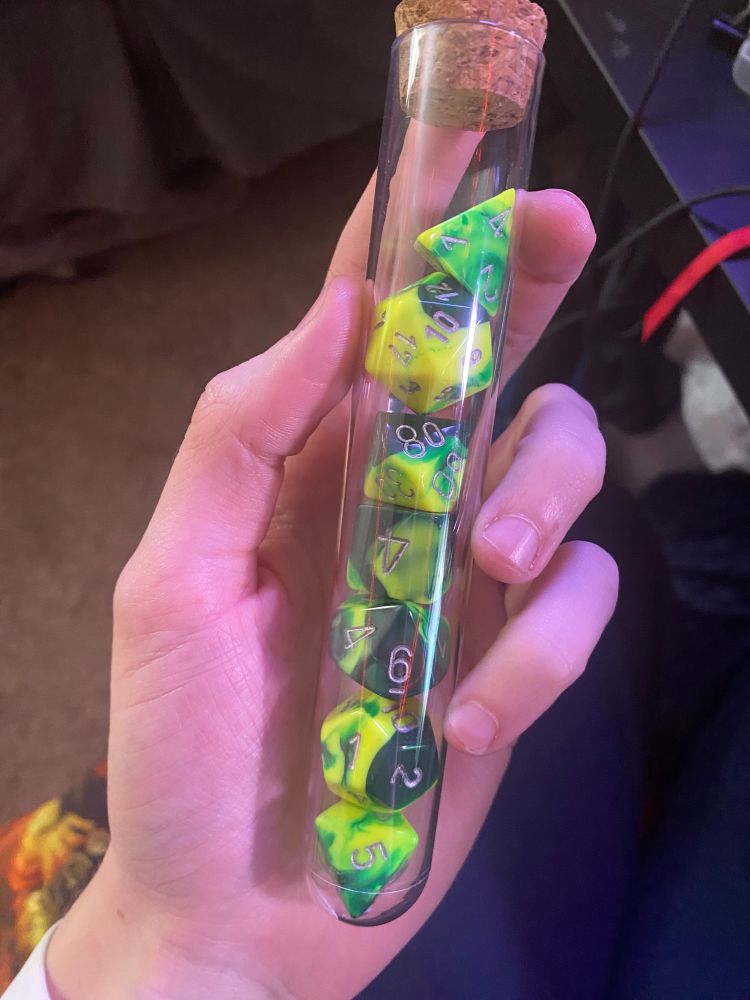 A hand holds a clear test tube vial, containing a set of dice. They are marbled dark green and almost electric yellow-green, with silver numbers