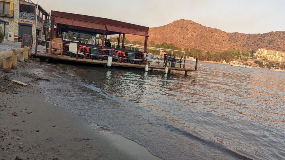 Seaside. On the left side, there's a deck that goes upon the sea. The deck has seats, tables and a roof. On the right side, there are mountains way back, and a hotel carved into them.