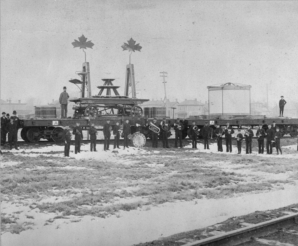 The Mammoth Cheese and its special wagon have been loaded onto flat cars at the Canadian Pacific Railway station in Perth on Saturday, April 15, 1893, and are ready to be coupled to the special World's Fair Cheese Train for transport to the Columbian Exhibition in Chicago the following Monday morning.  A marching band poses in front of the two railway cars. Note the two large sheet iron maple leaves decorating the wagon. Library and Archives of Canada photo.