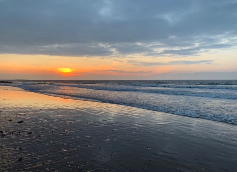 A sunset at high tide at a beach. The waves were calm, and the sky glowing golden orange as the sun set with some grey clouds above. The sky reflected on shallowest coating of water left on the sand just as the waves receded.