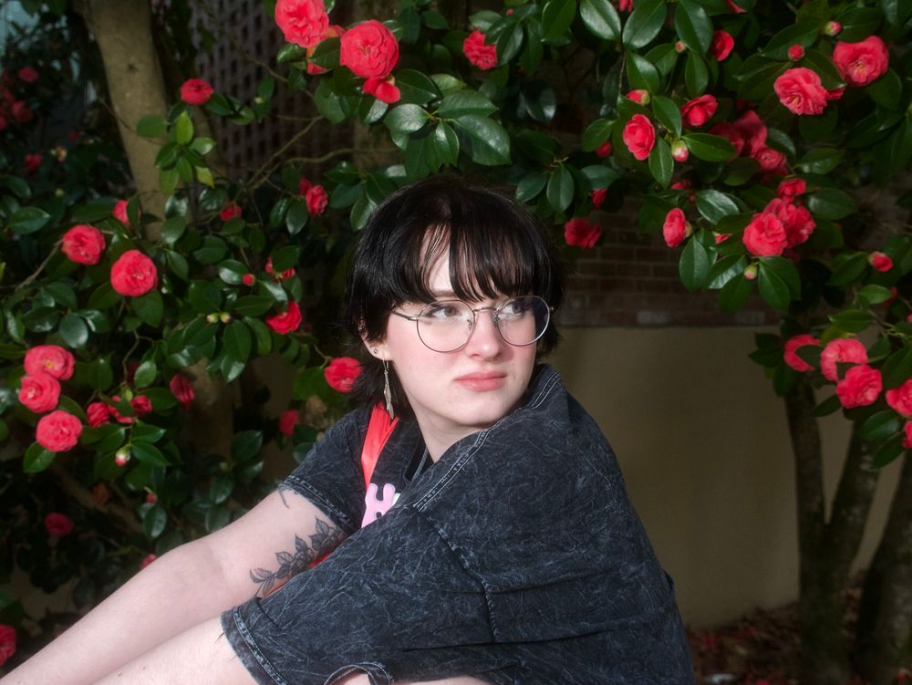 A woman in a black T-shirt with harsh lighting in front of a flower bush.
