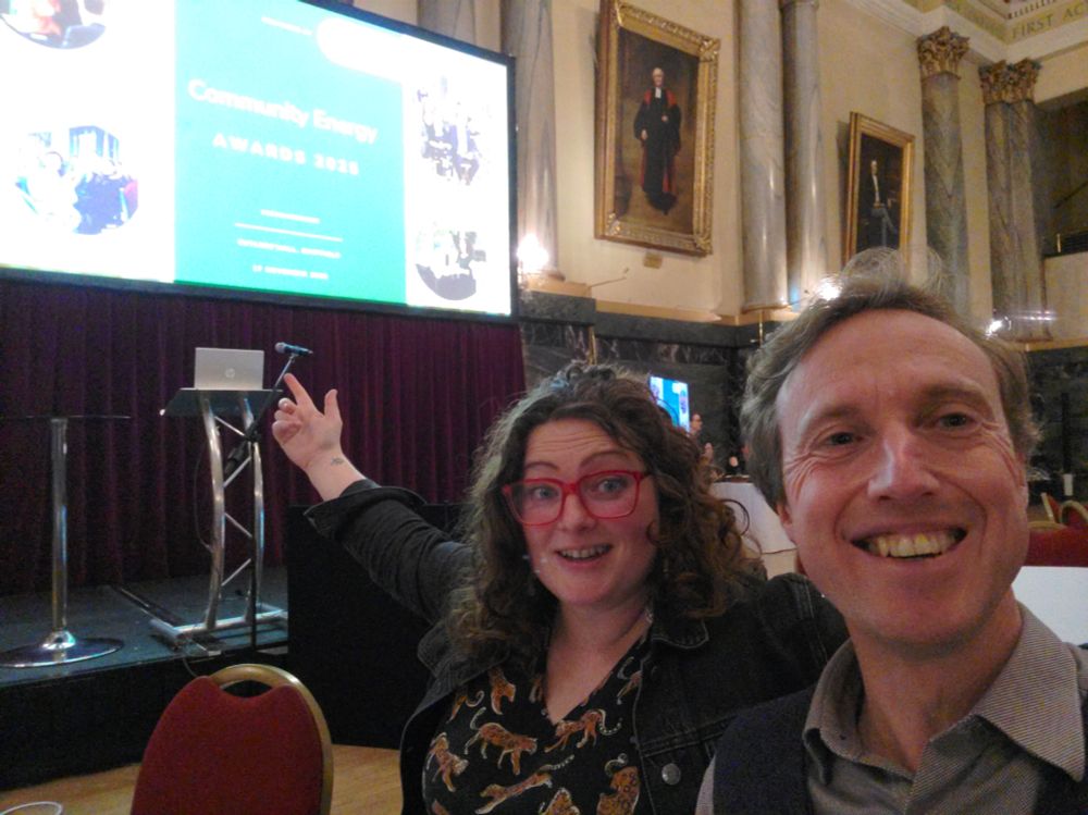 Slightly awkward selfie of two people (me and Abi from York Community Energy) awaiting the start of the Community Energy Awards 2025, indicated on a large screen to which Abi is pointing, in a big venerable hall (Cutlers' Hall in Sheffield) which is all columns and marble and hung with portraits of rich white men of past ages.