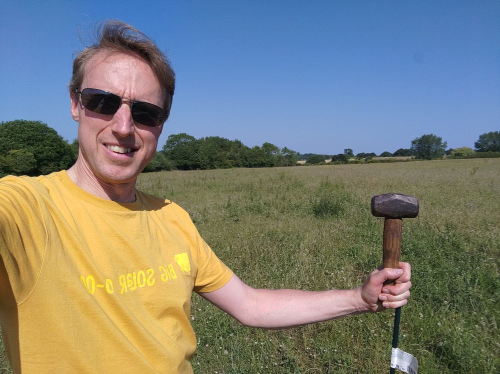 Fella standing in a very sunny field wearing dark glasses, a "Big Solar Co-op" T-shirt (barely legible cos the phone is on a stupid default setting to mirror the selfie camera) and holding a lumphammer in his left hand, which is resting on a stake he has just hammered in to the ground.