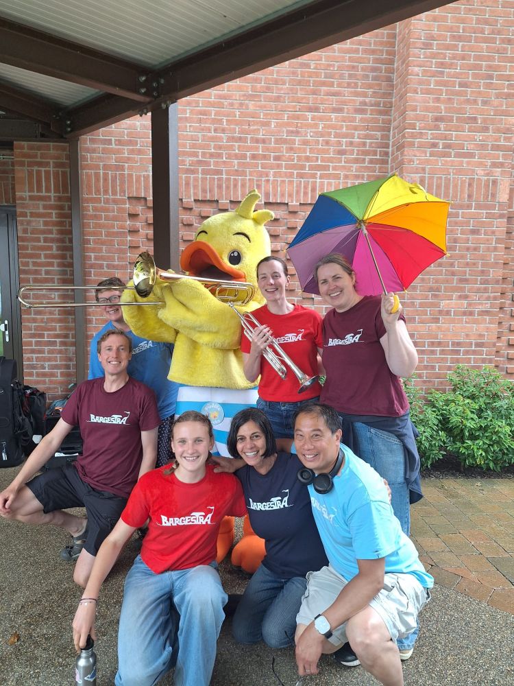 A group of people wearing 'Bargestra' t-shirts standing outside in a paved courtyard around a person in a duck mascot costume. One is holding a trumpet, another a colourful umbrella. The duck is holding a trombone.