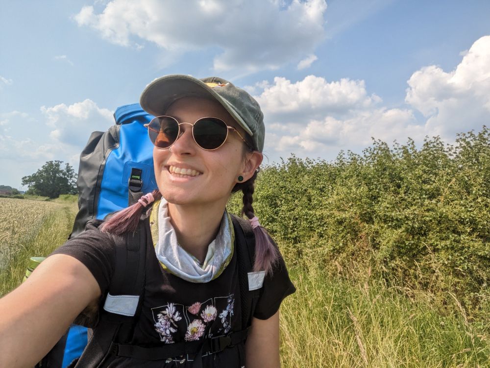 Picture of Natalie with a tree climbing kit in a field. She is wearing sunglasses and a cap. 