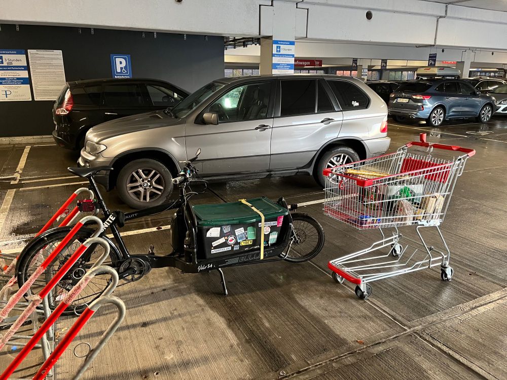 Mein Lastenrad auf einem Supermarktparkplatz. Davor ein Einkaufswagen. Daneben ein SUV, der genauso viel Platz einnimmt, wie insgesamt für Fahrräder zur Verfügung steht.