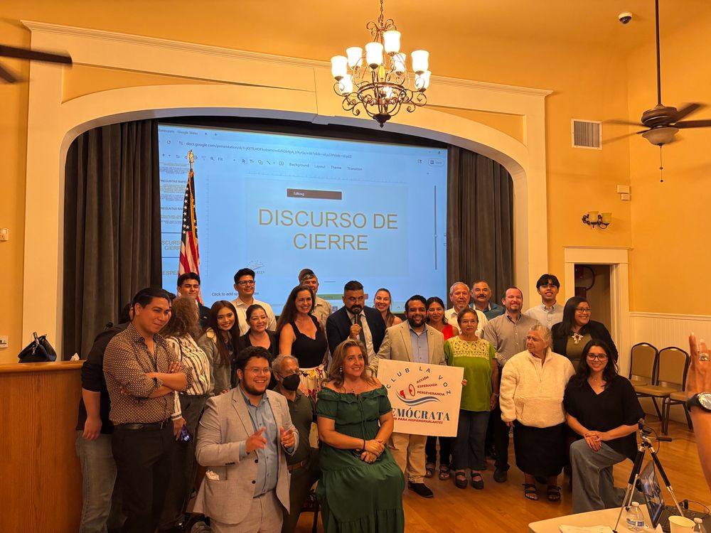 Group of people in front of a stage at a hall holding a sign that says Club La Voz Demócrata