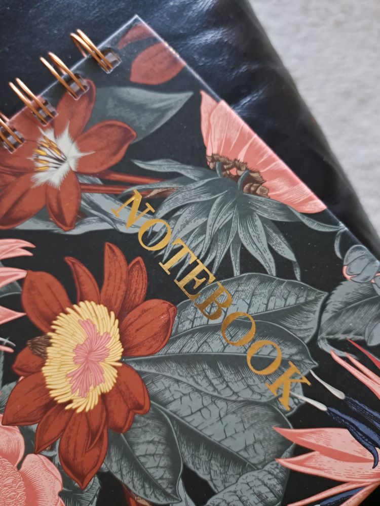 Op een zwarte leren bank met eronder een randje lichtgrijs kleed zie je de hoek van een kaft van een notitieboek. Het zit in elkaar met koperkleurige ringband en op de kaft staan rood-roze-oranje bloemen met grijze bladeren en in gouden letters het woord NOTEBOOK. 