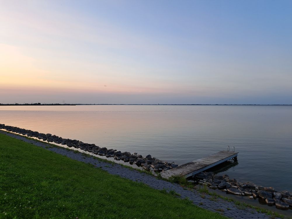 Zicht op het Markermeer vanaf de dijk bij Warder vlak voor zonsondergang. Links onder een schuin stuk groene dijk met onderaan grijze steenblokken. Na een kleine ondiepe geul ligt daar nog een minidijkje van grote blokken steen die onregelmatig op elkaar gelegd zijn. Rechts onder steekt een steiger het water in. Het water is vrij rustig en verkleurd in het licht van de ondergaande zon. Rechtsonder is het blauwgrijs, linksboven lichtoranje/roze met een blauwgrijze streep erin. De lucht boven het water verkleurt ook van lichtblauw rechtsboven tot oranje-roze aan de linkerkant. Aan de horizon is nog een streepje land te zien. 