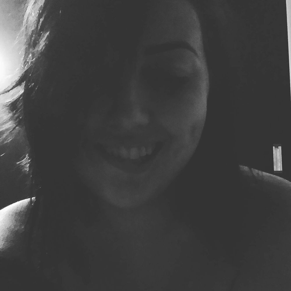 foto em escala de cinza, na sombra, com um ponto de luz vindo do canto superior esquerdo, por trás da minha cabeça. é um close do meu rosto e ombros nus. sou uma mulher branca de cabelos pretos, que na foto estão lisos, na altura dos ombros, repicados e partidos de lado. minhas sobrancelhas são escuras e bem definidas, meus olhos estão semicerrados, estou sorrindo e mordendo meu lábio inferior. em uma das bochechas aparece uma covinha bem marcada, a outra está coberta pelo meu cabelo.