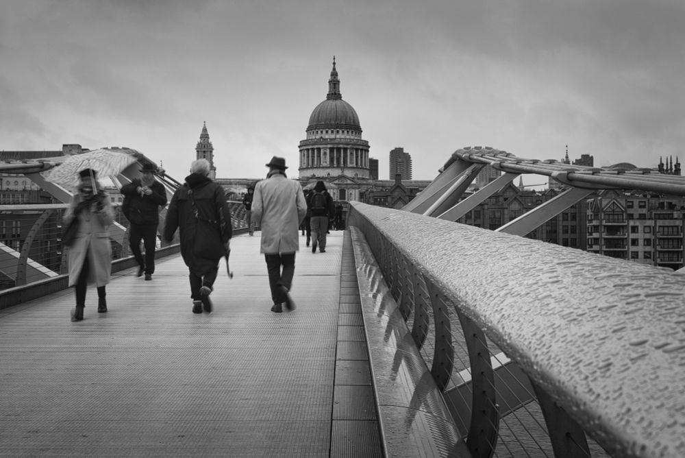 Taken from the middle of the Millennium Bridge over the River Thames. I was leaning on the rail on the right and raindrops can be seen on the surface. St Paul's cathedral is in the mid-distance. A long shutter speed has slightly blurred passing pedestrians. The image is in black and white