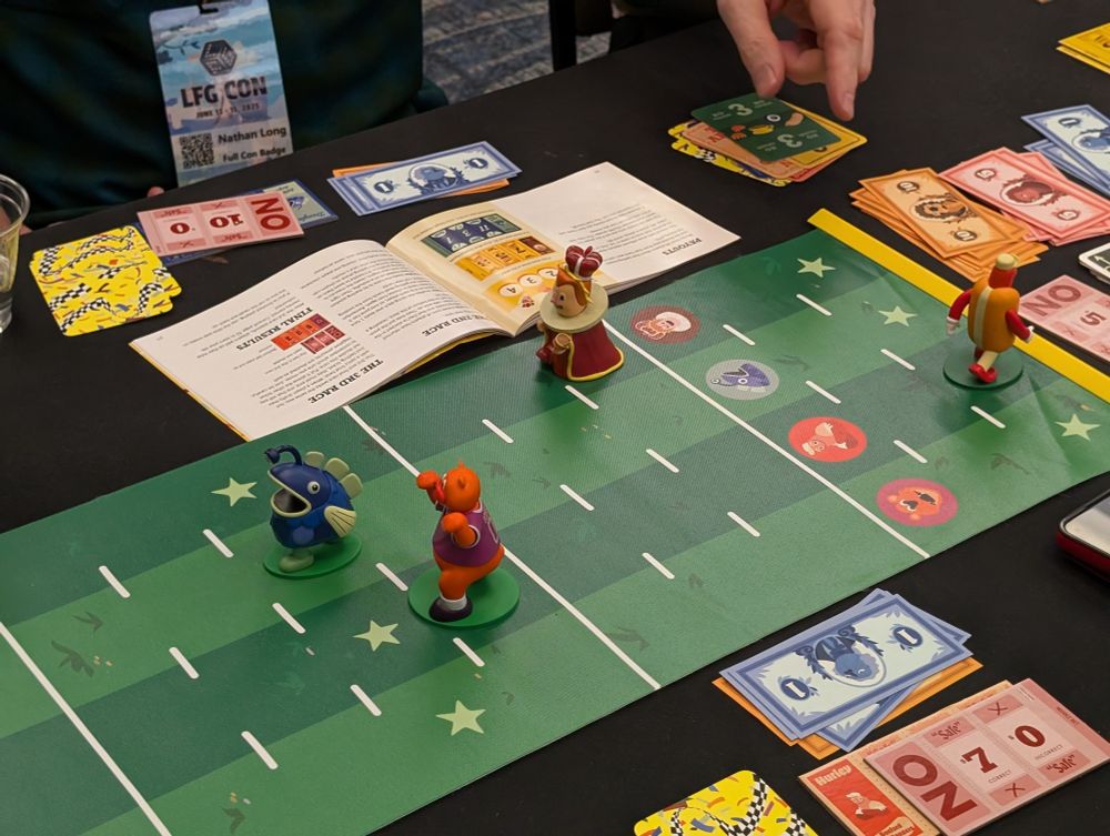Four different mascots racing along a grassy track, with the hot dog racer turned around running backwards. Stacks of cash and bets are off to the side.