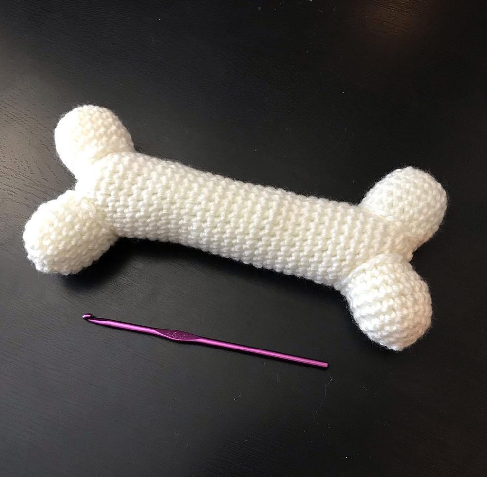 A stuffed, white, crocheted “bone”, similar to a dog bone treat in shape, next to a pink crochet hook.