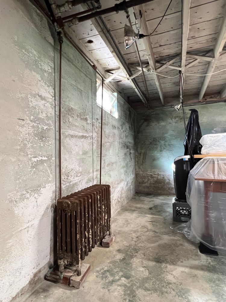 Unfinished basement. Rusty radiator on clay bricks. Joists. Concrete walls showing the remnants of a mural from the 1940s or 1950s and years of effervescence. #artyear #artstudio #artsky #elbowgrease #limewash