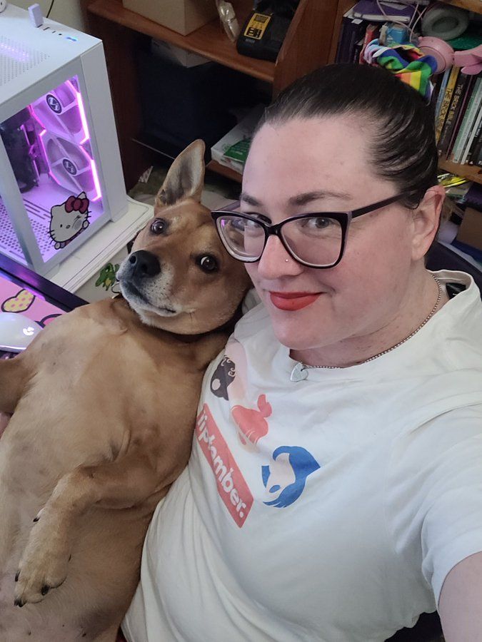  A person with their hair in a ponytail and wearing glasses is taking a selfie with their dog. The person is wearing a white T-shirt with a 