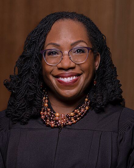 Supreme Court Justice Ketanji Brown Jackson smiling in judicial robes, a beaded necklace and glasses.