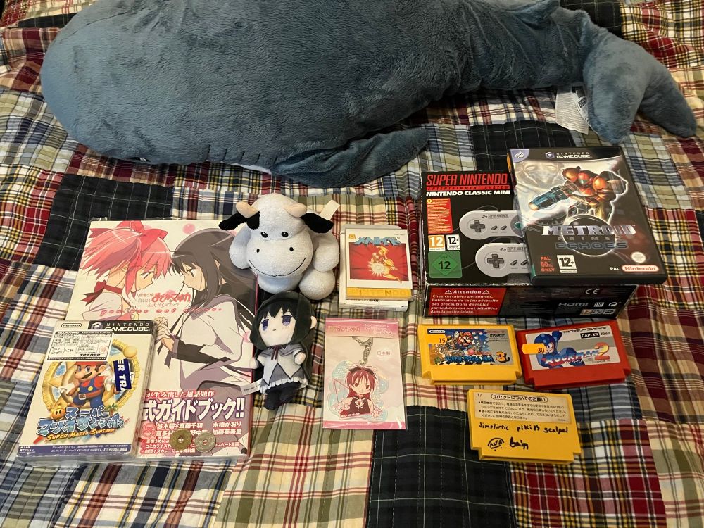 Souvenirs from my trip to Madrid, many of which are gifts from Aryll. Shown items are a BLÅHAJ shark plushie, Madoka Magica Guidebook "You Are Not Alone", Japanese Super Mario Sunshine, Cow toy, Homura Akemi keychain, FDS Metroid keychain, Kyoko Sakura keychain, CIB European SNES Mini, PAL Metroid Prime 2, Japanese Super Mario Bros. 3, Rockman 2, and my personal Famicom Super Mario Bros, cartridge signed by everyone who went on this trip