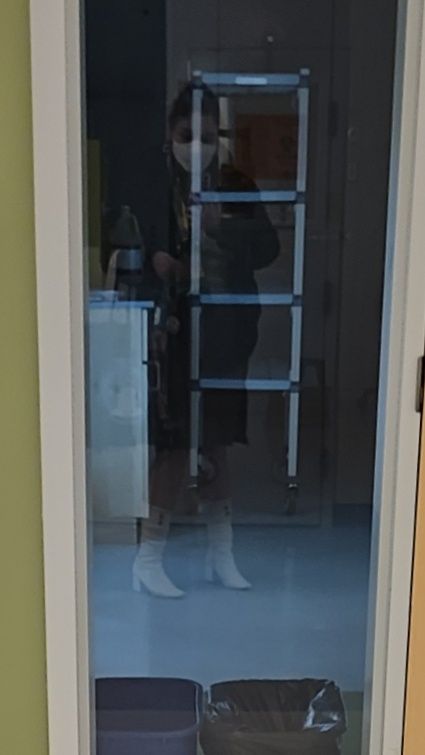 Selfie of my (person with hair up, wearing a black coat, skirt, white mask, and white boots) reflection in the window to an unoccupied room